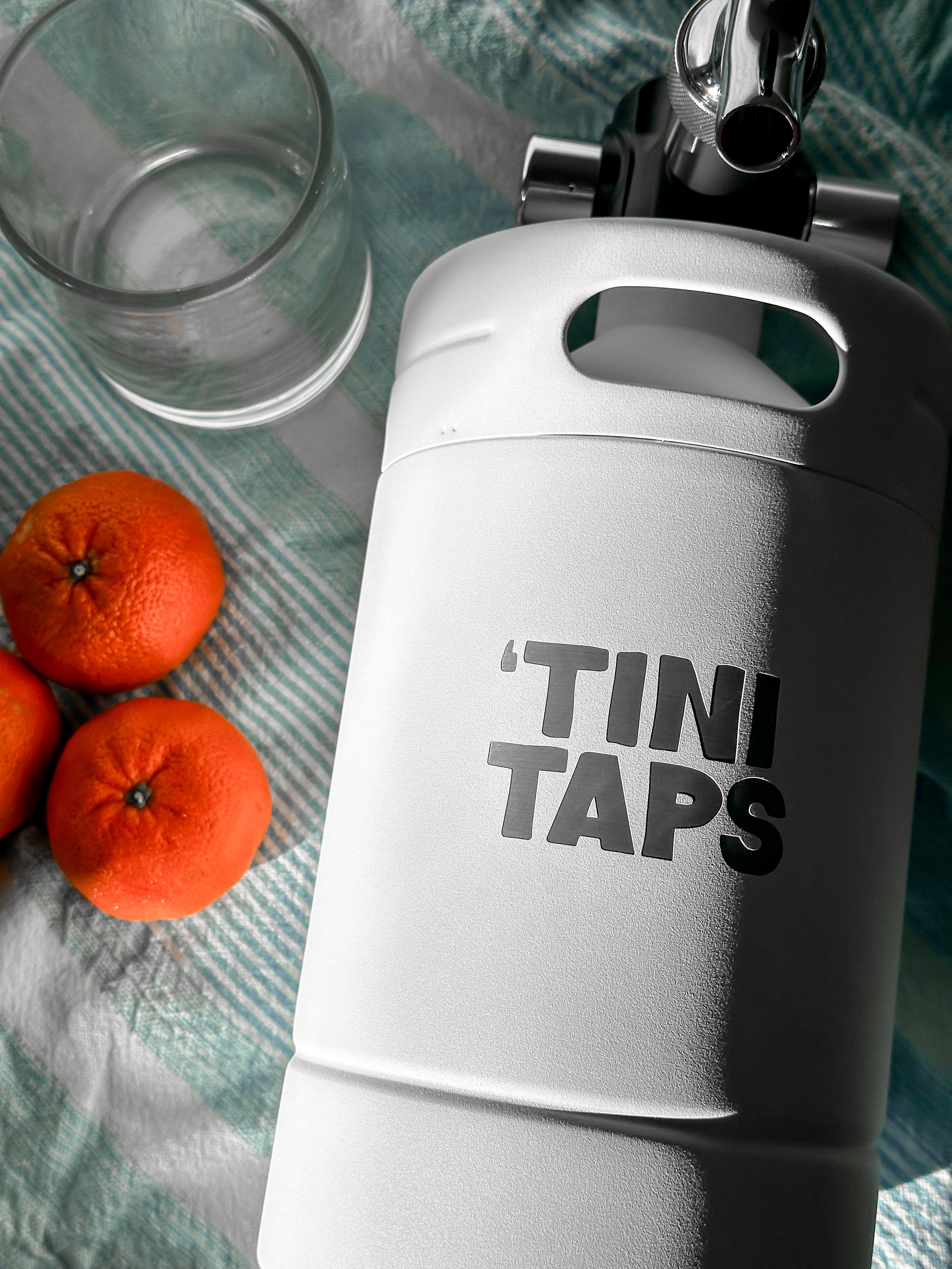 White 'TINI TAPS' keg with oranges and a glass on a textured surface